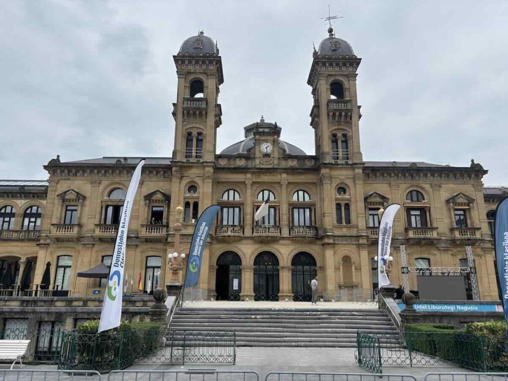 A symmetrical sandstone civic building with two domed towers and a central clock, wide steps leading up to arched doors, banners either side.