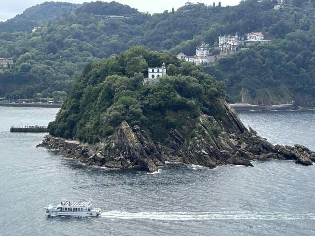 A steep wooded island with a white building near its summit and layered rock at the waterline, a small passenger ferry crossing in front.