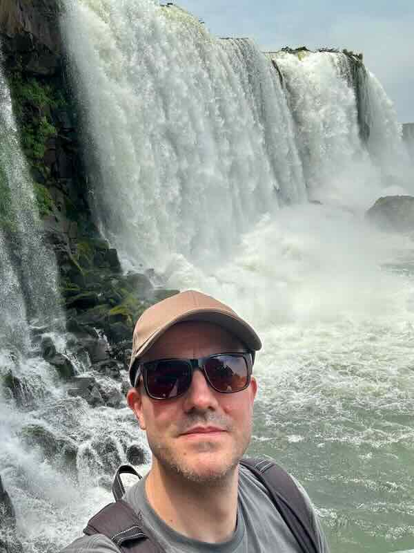 Chris in a cap and sunglasses in front of a wide curtain of falling water and churning white river below.
