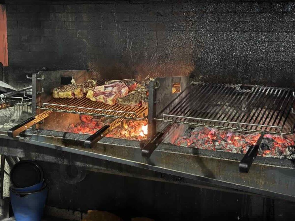 Thick bone-in steaks searing on a raised metal grill rack above glowing orange coals, soot-blackened wall behind.