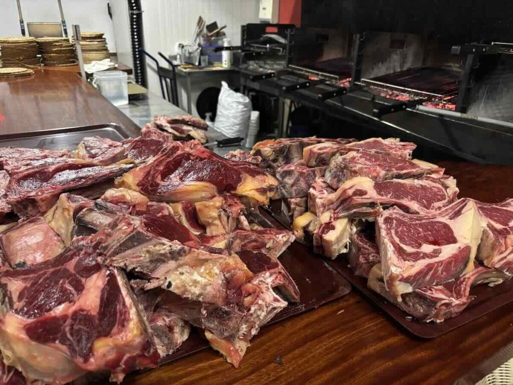 Two trays piled with raw bone-in ribeye steaks on a wooden counter, with the charcoal grill and glowing coals behind.