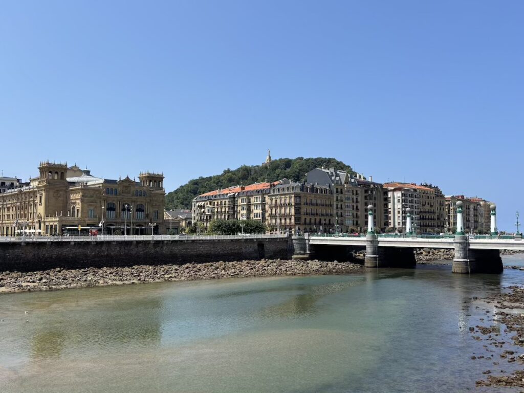 A road bridge with green and white lamp columns crossing shallow green water at low tide, grand buildings along the far bank and a wooded hill with a hilltop statue behind.