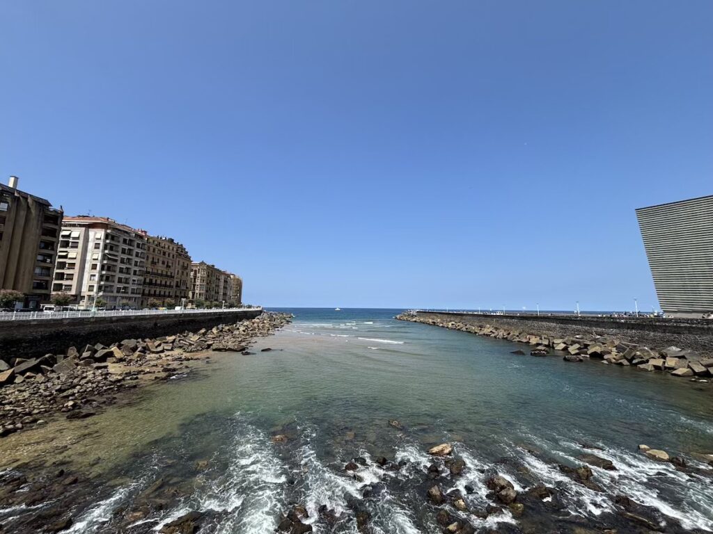 Shallow water flowing over rocks between two boulder breakwaters out to open sea, apartment blocks on the left and a ridged glass facade on the right.