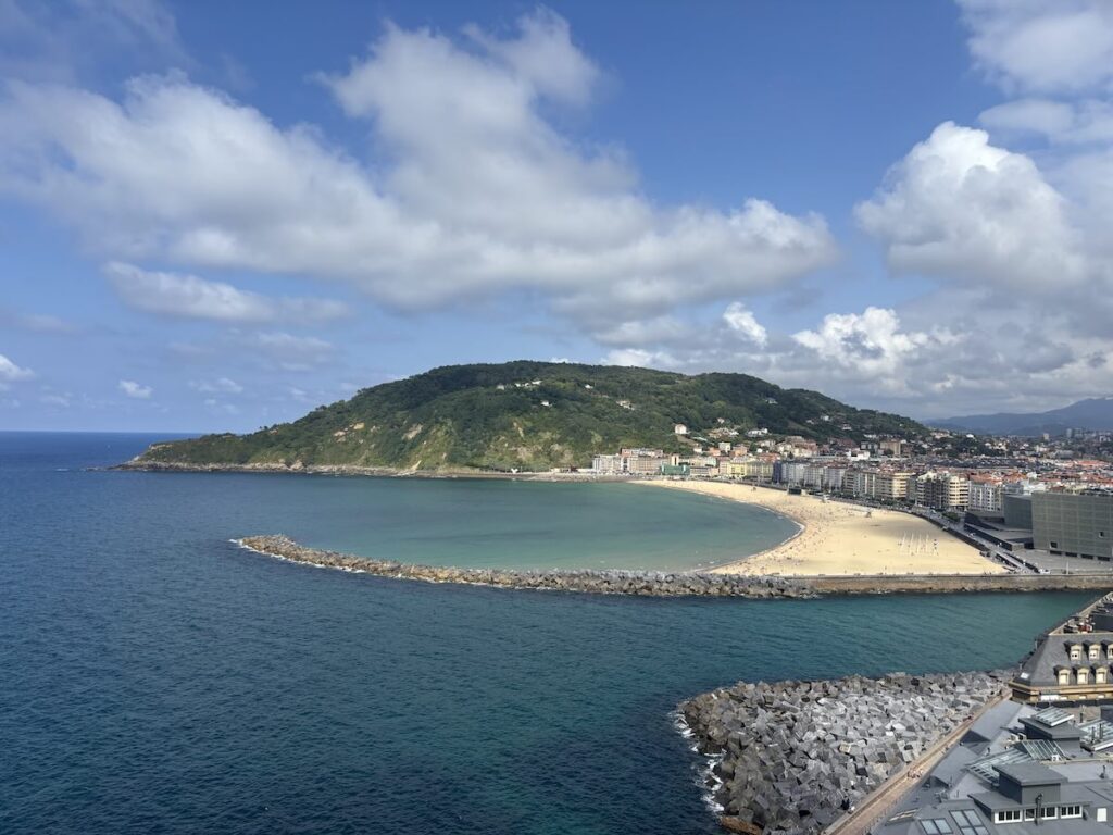A broad sand beach behind a boulder breakwater with a green headland rising beyond it, dark blue sea in the foreground and buildings along the shore.