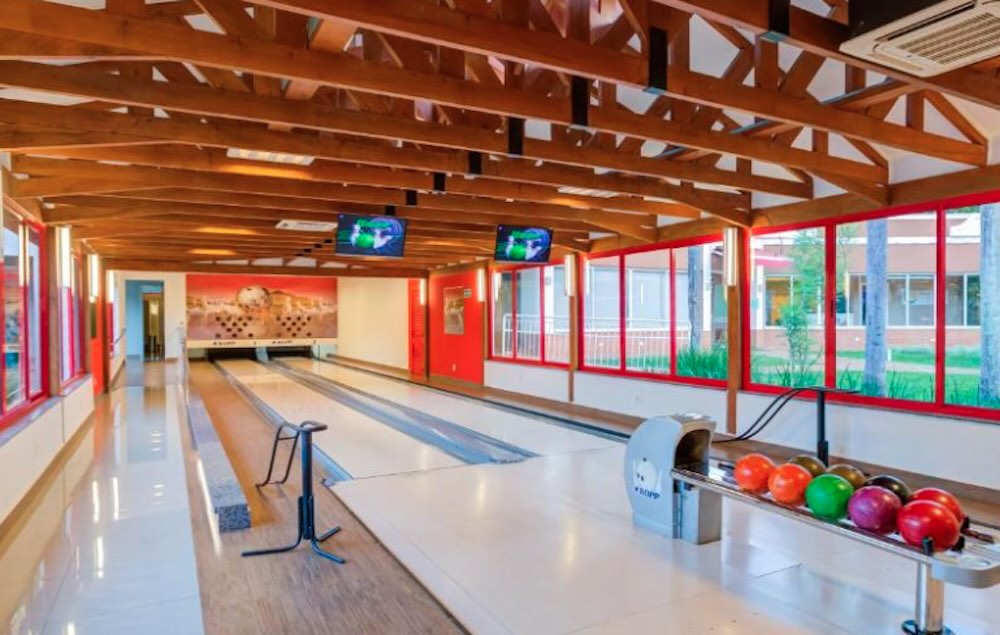 Two-lane bowling alley under a wooden beamed roof, with red window frames and coloured balls racked by the return
