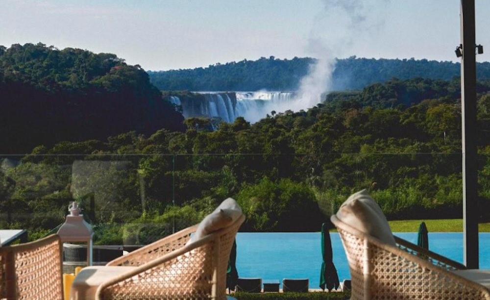 View from a poolside terrace over a blue pool and lawn to the falls and their rising spray across the gorge beyond