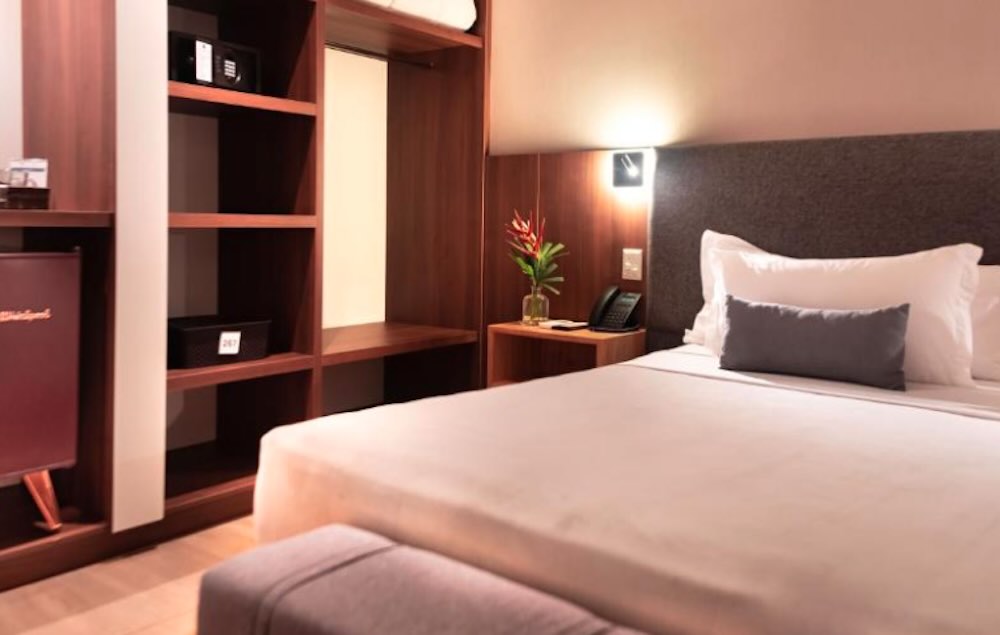Hotel room with dark wood shelving and an open wardrobe, a large bed in white linen and a single flower on the bedside table