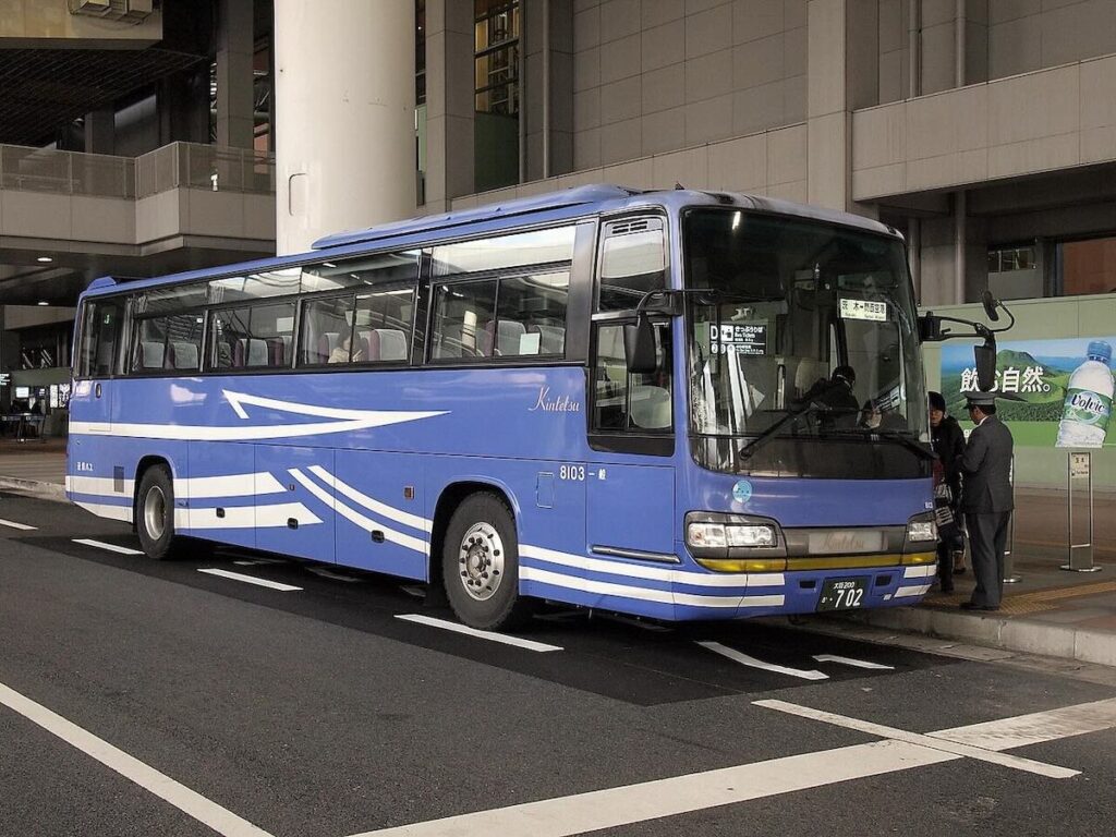 A blue and white Kintetsu airport limousine bus at a terminal stop after dark, with a uniformed attendant helping a passenger board.