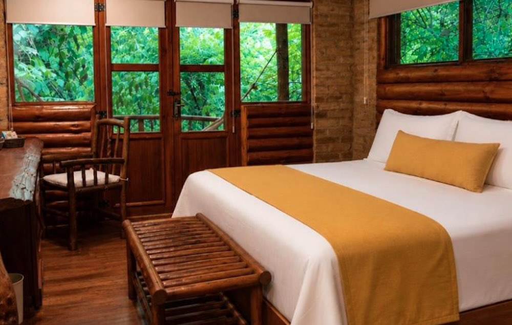 Log cabin bedroom with a mustard bed runner and wooden doors opening straight onto dense green foliage outside