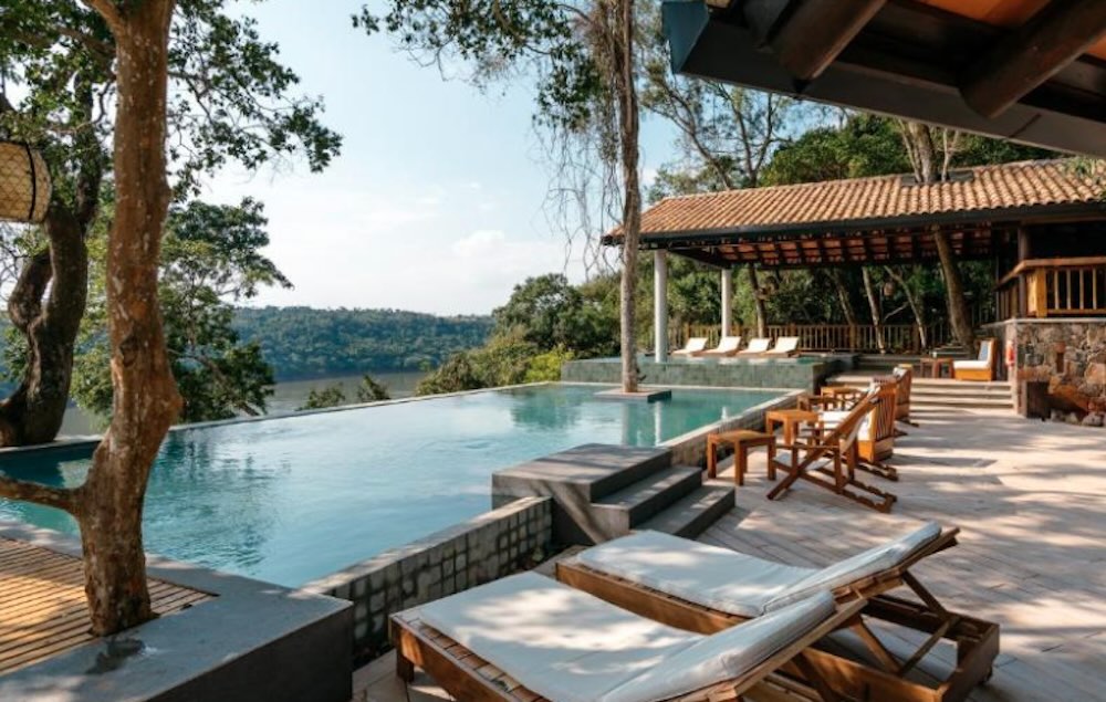 Infinity pool on a timber deck with sun loungers, framed by tall trees and looking out over water and a forested hillside
