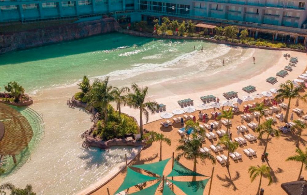 Aerial view of a resort wave pool with its own sand beach, rows of white loungers, palm trees and green shade sails