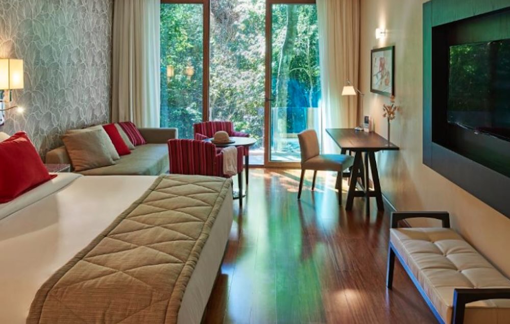 Spacious room with a sofa, desk and wall-mounted TV, and floor-to-ceiling glass doors opening onto the forest