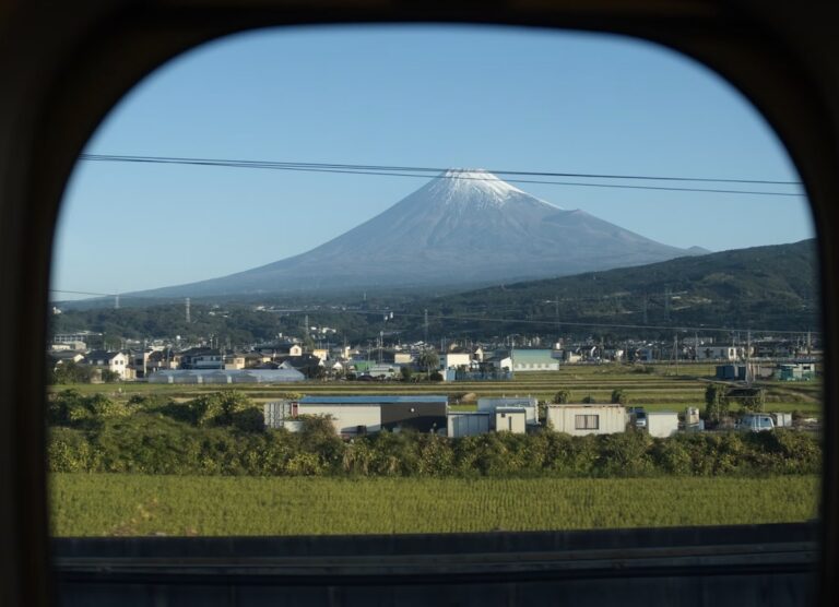 Mount Fuji framed by a shinkansen window on a clear morning, snow on the summit above rice fields and low houses.