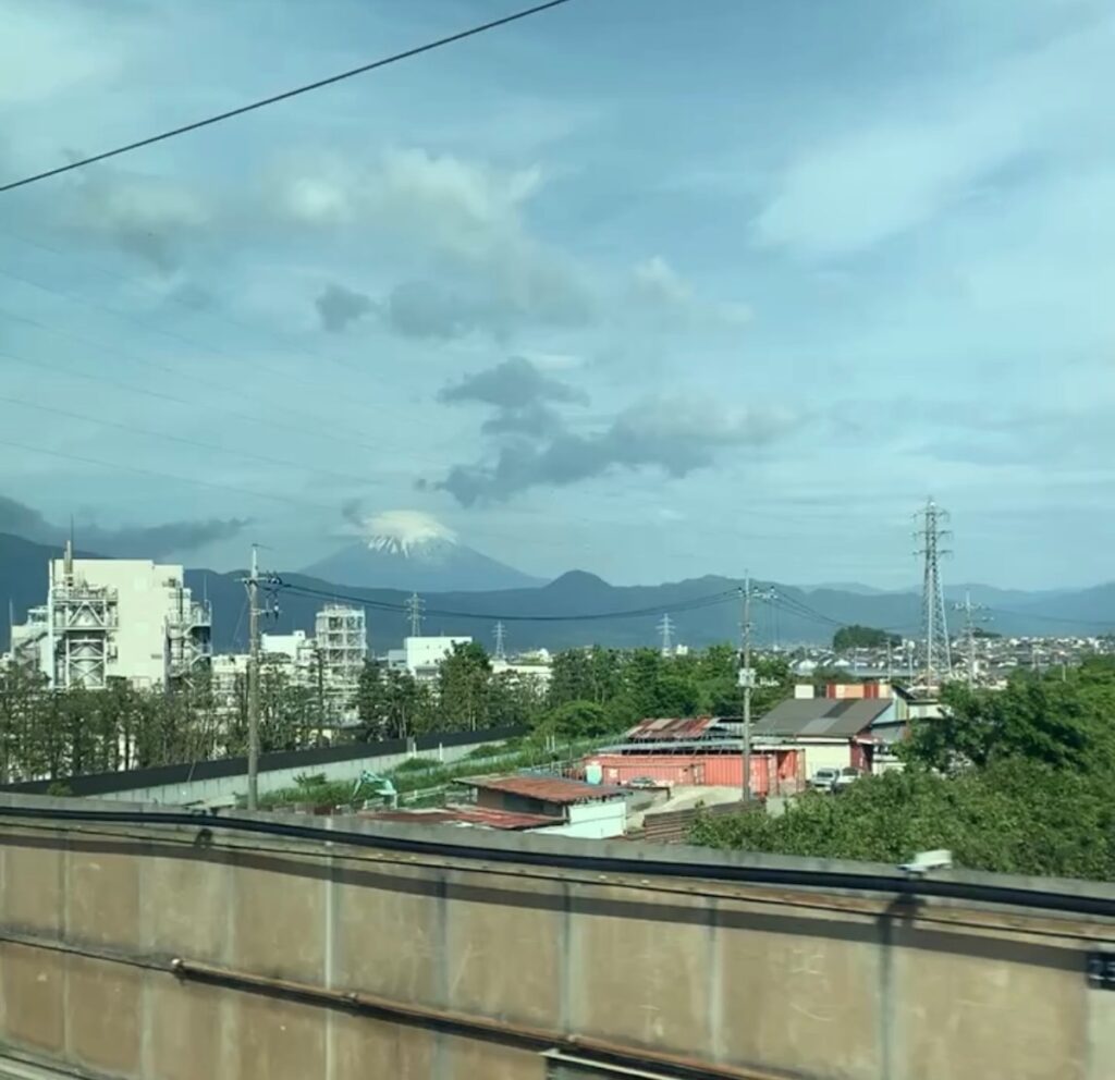 Mount Fuji with its summit in cloud, seen past factories and power lines from a Tokyo to Osaka bullet train window.