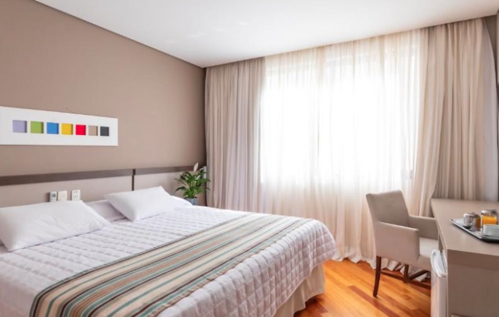 Double room with a striped bed runner, pale wood floor and an armchair beside a desk at a bright curtained window