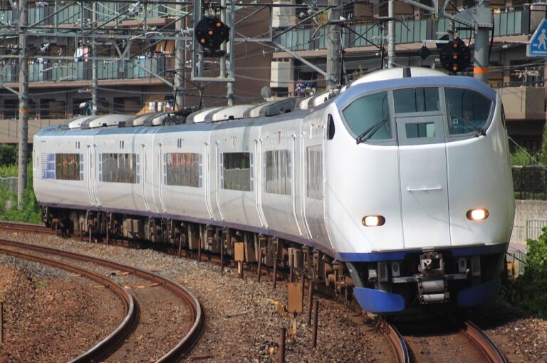 The white and blue Haruka limited express train rounding a curve on the approach to a station, with overhead lines above the track.