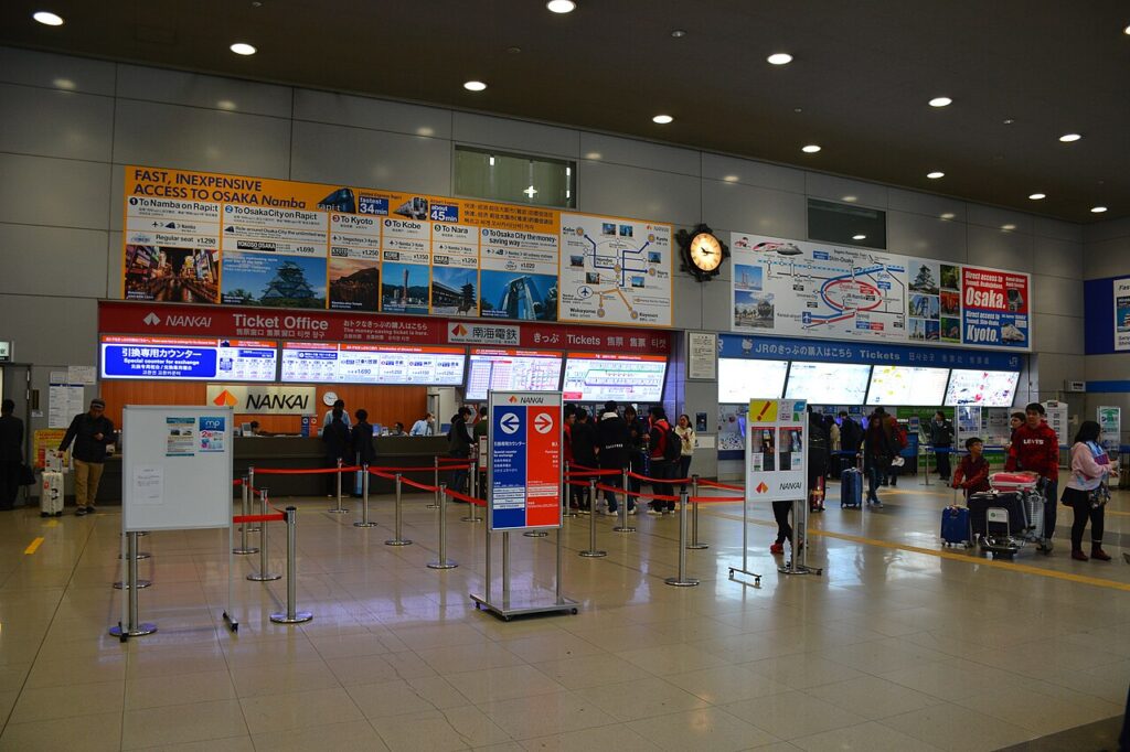The red Nankai ticket office and the blue JR ticket counters side by side in the concourse at Kansai Airport Station, with route boards above.