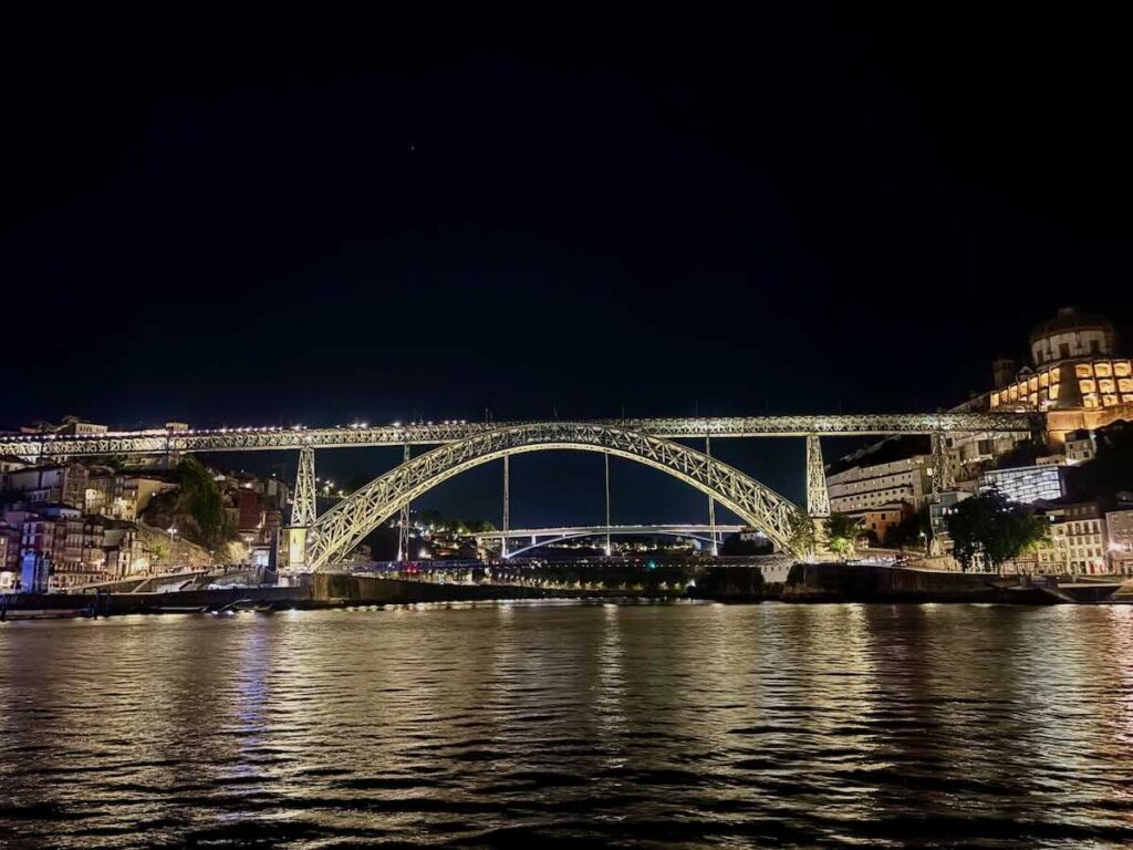 Dom Luís I Bridge in Porto lit up at night, its iron arch reflected in the Douro River with Ribeira and Gaia either side