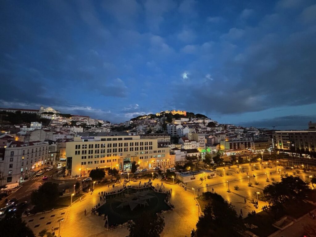 Martim Moniz square in Lisbon lit up at dusk from a rooftop, with São Jorge Castle floodlit on the hill behind